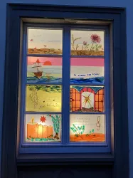 Ein Fenster mit bunten, handgemalten Bildern zeigt verschiedene Landschaften und Motive. Die Kunstwerke sind lebhaft und fröhlich, mit einem maritimen Thema und dekorativen Elementen.