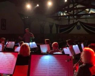 Ein Musiker leitet ein Orchester in einer gut beleuchteten Halle. Die Musiker sitzen mit Notenblättern vor sich und konzentrieren sich auf die Aufführung.