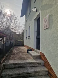 Ein einfacher Hauseingang mit Steinstufen und einer weißen Wand. Die Sonne scheint leicht hinter den Bäumen.