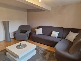 Ein gemütliches Wohnzimmer mit einem grauen Sofa und zwei Sesseln. Ein moderner Couchtisch steht in der Mitte auf einem hellen Teppich.