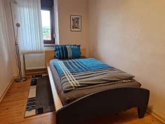 Ein einfaches Schlafzimmer mit einem einzelnen Bett und bunten Kissen. Das Zimmer hat Holzfußboden und ein großes Fenster mit Vorhängen.