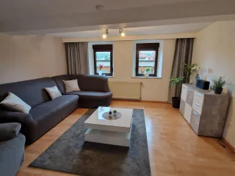 Ein modernes Wohnzimmer mit einer grauen Couch und einem Couchtisch. Große Fenster lassen viel Licht herein, und es gibt Pflanzen und Dekorationen im Raum.