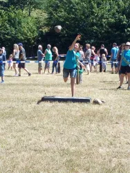Eine Person in einem blauen T-Shirt wirft einen Stein auf einer Wiese. Im Hintergrund sind viele Zuschauer und ein sonniger Himmel zu sehen.