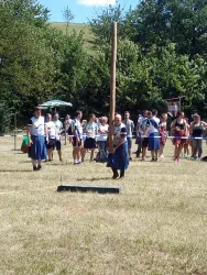 Eine Gruppe von Menschen beobachtet einen traditionellen Wettkampf im Freien. Ein Teilnehmer steht neben einem hohen Holzstock und trägt eine Tracht.