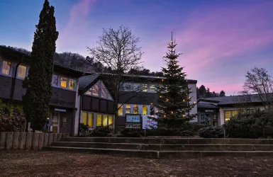 Ein Schulgebäude mit beleuchtetem Weihnachtsbaum im Vordergrund. Der Himmel zeigt während der Dämmerung schöne pinke und lila Farbtöne.