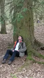 Eine Person sitzt entspannt unter einem Baum im Wald. Der Boden ist mit Laub bedeckt und die Umgebung ist grün und ruhig.