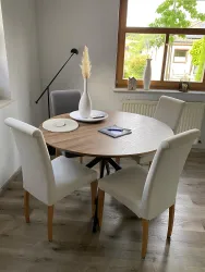 Ein runder Holztisch mit zwei weißen Stühlen und einem grauen Stuhl. Auf dem Tisch stehen eine Vase und ein Tablet, während das Licht durch ein Fenster fällt.