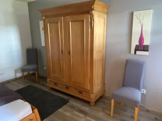 Ein gemütliches Schlafzimmer mit einem großen Holzschrank und zwei grauen Stühlen. Der Boden ist aus Holz und es gibt ein modernes Wandbild.
