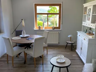 Ein einladendes Wohnzimmer mit einem runden Tisch und weißen Stühlen. Durch das Fenster scheint das Tageslicht und bietet einen schönen Blick ins Grüne.