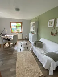 Ein helles Wohnzimmer mit einer gemütlichen Couch und einem Tisch. An der Wand befinden sich Bilder und eine Dekoration, und der Raum hat große Fenster, die Tageslicht hereinkommen lassen.