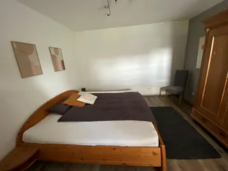 Ein helles, modernes Schlafzimmer mit einem großen Holzbett. An der Wand hängen zwei einfache Bilder und ein Sessel steht in der Ecke.