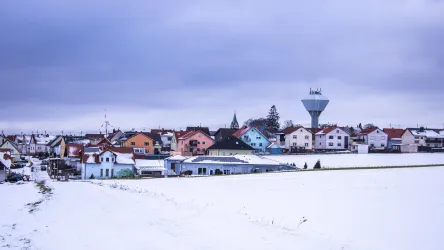 Eine verschneite Landschaft mit einem kleinen Dorf. Die Häuser sind bunt bemalt und ein Wasserturm ist im Hintergrund sichtbar.
