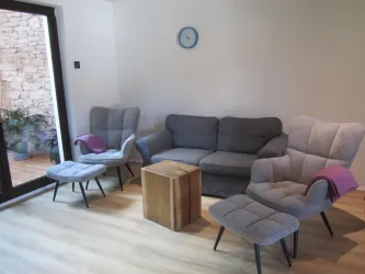 Ein gemütlicher Wohnraum mit einem grauen Sofa und zwei Sesseln. Ein quadratischer Tisch aus Holz steht in der Mitte, und eine Uhr hängt an der Wand.