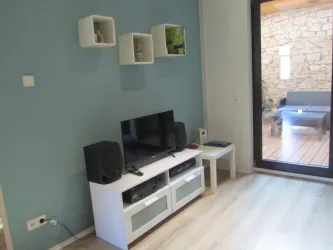 Ein moderner Wohnraum mit einem Fernseher auf einem weißen Medienzentrum und Lautsprechern. Der Raum hat helle Wände und einen Blick auf eine möblierte Terrasse.