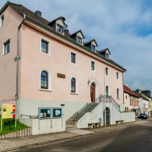 Ein pinkes Gebäude an einer ruhigen Straße mit Stufen und Fenstern. Neben dem Gebäude sind grüne Wiesen und weitere Häuser sichtbar.