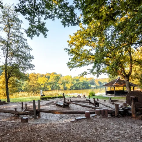 Ein ruhiger Park mit großen Bäumen und einem klaren See im Hintergrund. Im Vordergrund sind Holzspielgeräte und eine überdachte Sitzgelegenheit zu sehen.