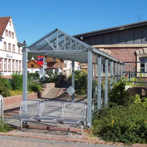 Eine moderne Bushaltestelle mit einem transparenten Dach und Sitzbänken. Im Hintergrund sieht man ein historisches Gebäude und fußläufige Wege.