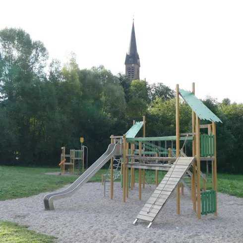 Ein Spielplatz mit Rutschen und Klettergerüsten steht in einem grünen Park. Im Hintergrund ist ein Kirchturm sichtbar.