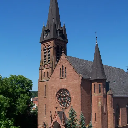 Eine schöne Kirche mit einem hohen Turm und roten Ziegeln. Umgeben von Bäumen und einem klaren blauen Himmel.