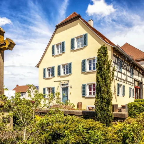 Ein charmantes Haus mit bunten Fensterläden und einem historischen Kreuz im Vordergrund. Die Landschaft ist grün und die Umgebung wirkt einladend.