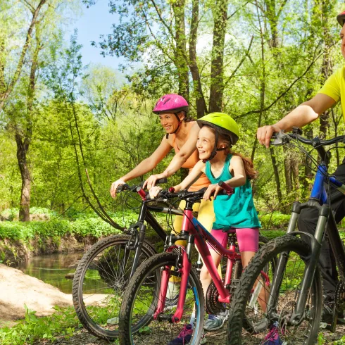 Eine Familie mit drei Radfahrern steht in der Natur. Sie tragen Helme und genießen die Zeit zusammen an einem kleinen Fluss.