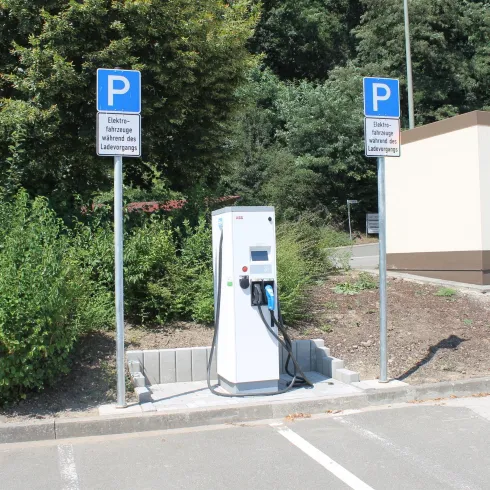 Eine Ladestation für Elektroautos steht auf einem Parkplatz. Im Hintergrund sind grüne Büsche und ein Gebäude sichtbar.