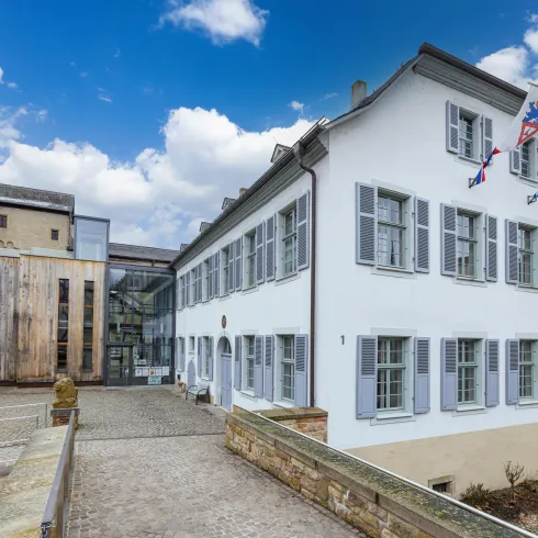 Ein modernes Gebäude mit traditioneller Architektur steht neben einer historischen Burg. Der Himmel ist bewölkt und es gibt eine ruhige Umgebung.
