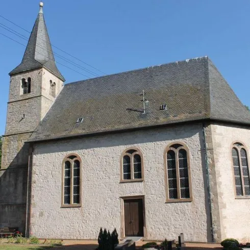 Eine alte Kirche mit einem hohen Turm und großen Fenstern. Die Fassade ist aus hellem Stein und die Umgebung ist grün und ruhig.
