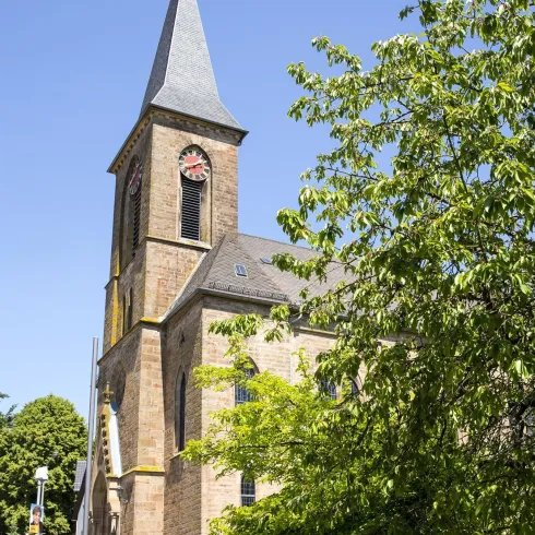 Eine alte Kirche mit einem hohen Turm und bunten Fenstern. Umgeben von grünen Bäumen und klarem Himmel.