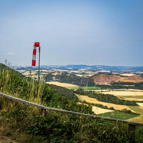 Ein weitläufiges Landschaftsbild mit sanften Hügeln und Feldern in verschiedenen Farbtönen. Ein Windsack weist auf die Windrichtung hin, vor einer wolkenlosen blauen Skyline.