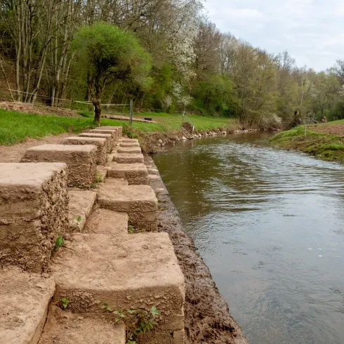 Ein ruhiger Fluss mit angrenzenden, grünen Wiesen. Am Ufer sind große Steine zu sehen, die als Stufen ausgelegt sind.