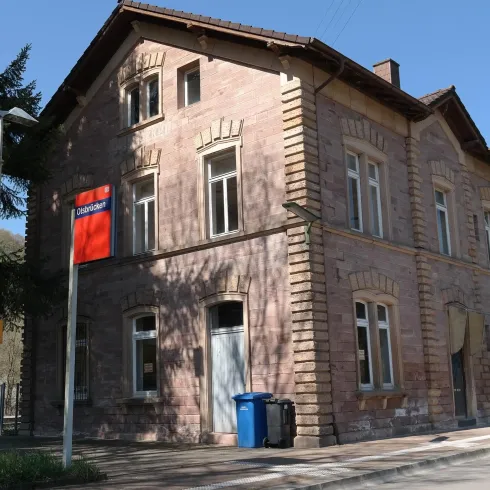 Ein altes Bahnhofsgebäude aus rotem Stein mit Fensterläden. Neben dem Gebäude stehen Mülltonnen und es ist ein klarer Himmel zu sehen.