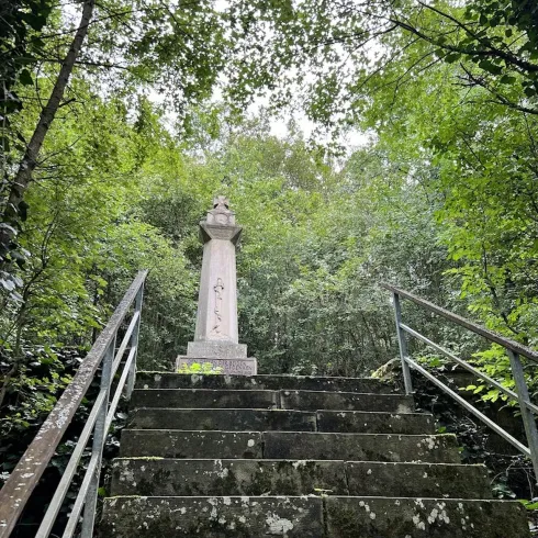 Eine steinerne Stufenanlage führt zu einem Denkmal, das von üppigem Grün umgeben ist. Die Atmosphäre ist ruhig und naturnah.