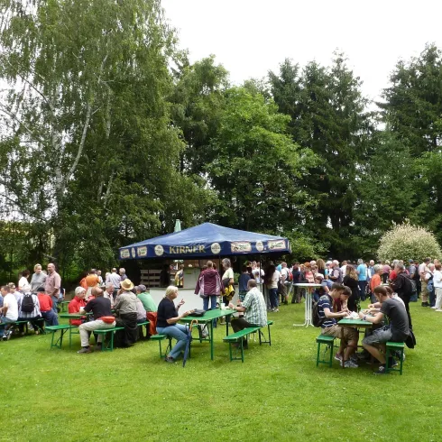 Eine große Menschenmenge versammelt sich auf einer Wiese mit Tischen und Bänken. Im Hintergrund sind Bäume und ein Zelt zu sehen.