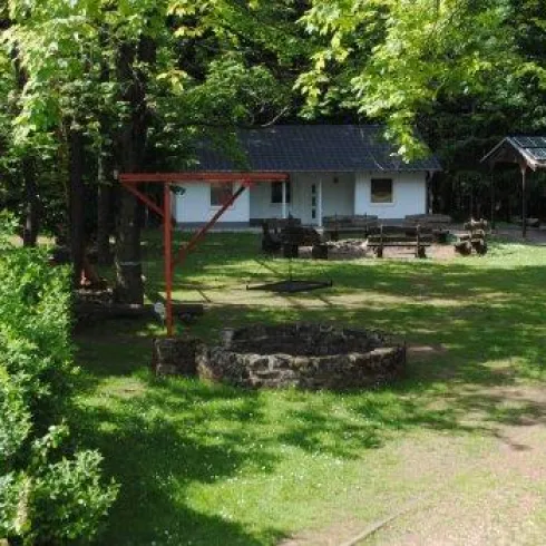 Eine ruhige Landschaft mit mehreren kleinen Gebäuden, umgeben von Bäumen und grünem Gras. In der Mitte befindet sich ein Lagerfeuerplatz.