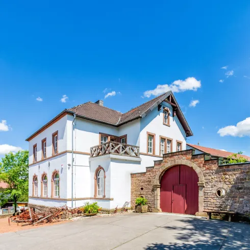 Ein schönes, historisches Gebäude mit einem großen Tor und einem gepflegten Garten. Der Himmel ist klar und blau, umgeben von grünen Bäumen.