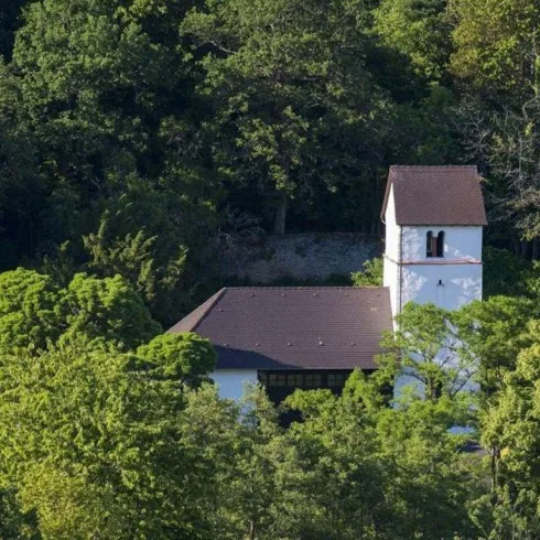 Eine kleine Kirche umgeben von dichtem Grün. Der Kirchturm ragt über die Bäume hinaus.