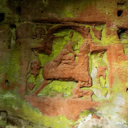Eine alte Felswand mit ausgestochenen Figuren und Moosbewuchs. Die Muster zeigen eine historische Darstellung, die in die Wand gemeißelt wurde.