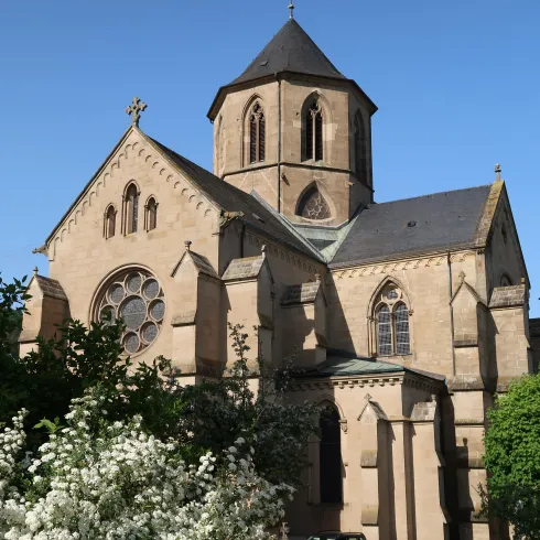 Eine schöne Kirche mit einem markanten Turm und großen Fenstern. Umgeben von grünen Pflanzen und blühenden Sträuchern.