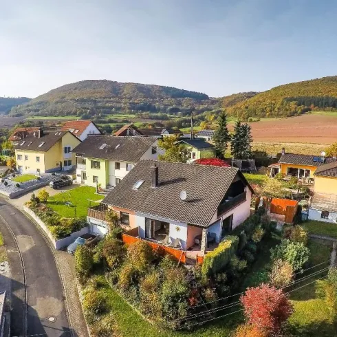 Eine malerische Wohngegend mit mehreren Häusern und grünem Garten. Im Hintergrund sind sanfte Hügel und ein klarer Himmel zu sehen.