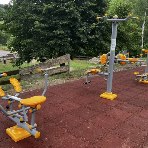 Ein Freiluft-Fitnessbereich mit mehreren Geräten in orange und grau. Der Boden ist mit roten Gummimatten ausgelegt und von grünen Bäumen umgeben.