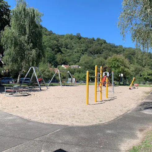 Ein Spielplatz mit Sandfläche und verschiedenen Spielgeräten steht in einem grünen Umfeld. Im Hintergrund sind Bäume und sanfte Hügel zu sehen.