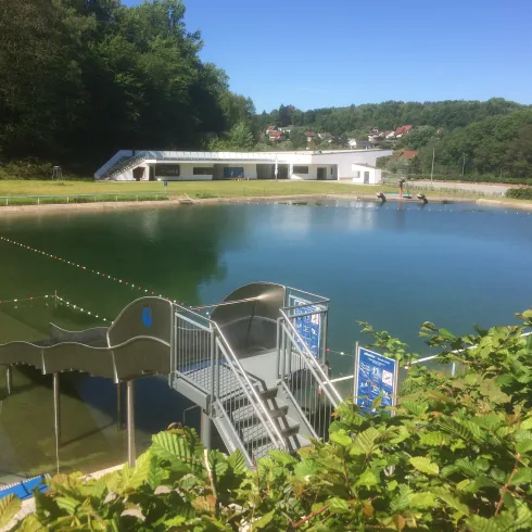 Ein ruhiger Badesee mit einem Zugangssteg und einer modernen Badanlage im Hintergrund. Umgeben von grüner Natur und einem klaren blauen Himmel.