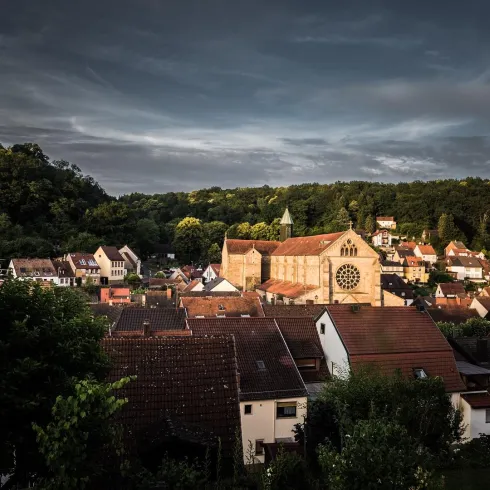 Eine malerische Stadt mit traditioneller Architektur und einer Kirche im Vordergrund. Im Hintergrund erheben sich grüne Hügel unter einem bewölkten Himmel.