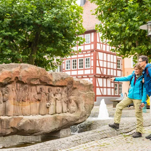 Ein Paar steht vor einem bunten Felsen mit Reliefs, während sie in einer malerischen Stadt spazieren. Im Hintergrund sind historische Gebäude und Bäume zu sehen.