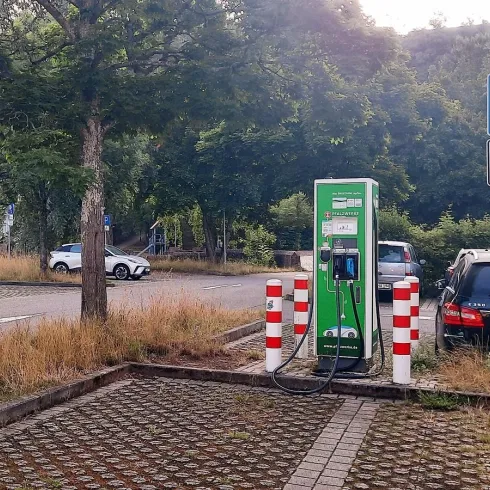 Eine Ladestation für Elektroautos in einer ruhigen Parkumgebung. Im Hintergrund sind parkende Autos und Bäume sichtbar.