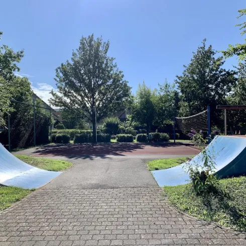 Ein Skatepark mit zwei blauen Rampen und viel Grün umgeben. Der Himmel ist klar und sonnig.