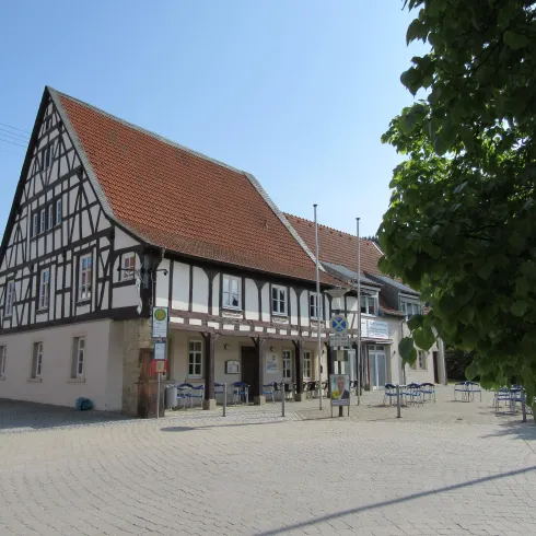 Ein traditionelles Fachwerkhaus mit einem roten Satteldach. Vor dem Gebäude sind Sitzgelegenheiten im Freien und es gibt einen klaren blauen Himmel.