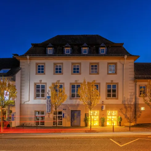 Ein historisches Gebäude mit einem hellen Fassaden und Bäumen im Vordergrund. Die Szene ist bei Nacht mit einem klaren, blauen Himmel.