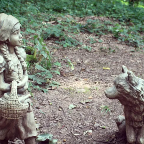 Eine Statue von Rotkäppchen mit einem Korb steht in einem Wald. Vor ihr sitzt ein Wolf.